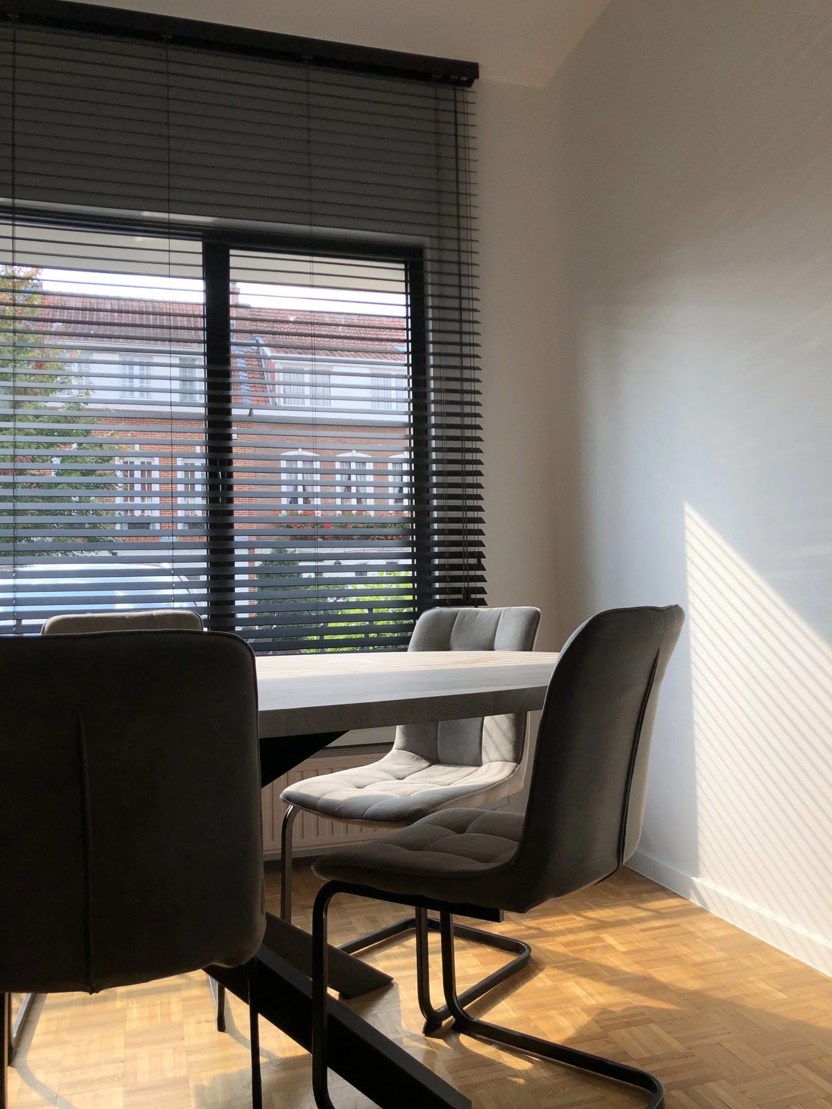 A dining room with a window covered by black blinds, a white table, and four beige cushioned chairs with metal legs.