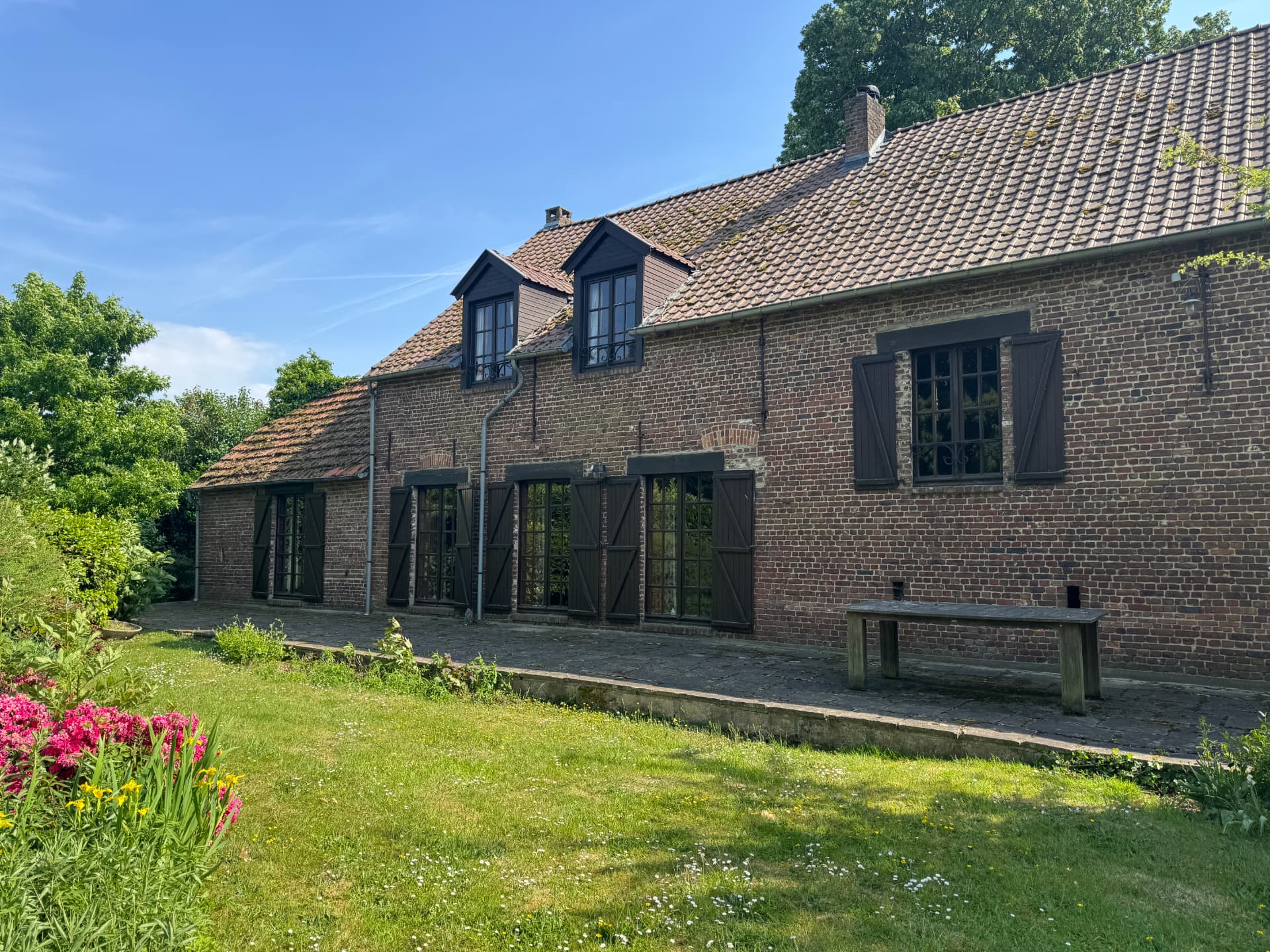 Behind the scenes: langgevel van de bestaande hoeve met houten luiken en tuin voor de renovatie.