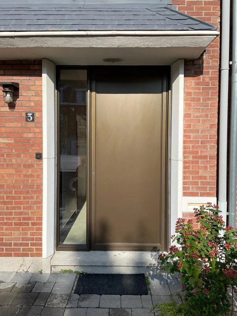 A brick house entrance with a modern metal front door, a small window beside it, a black outdoor light fixture and a flowering bush in front.